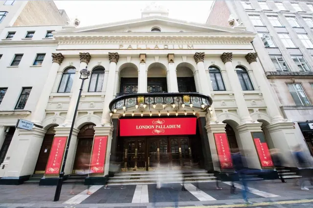 The front of the London Palladium theatre with red banners, a columned entrance and blurred pedestrians crossing the street.