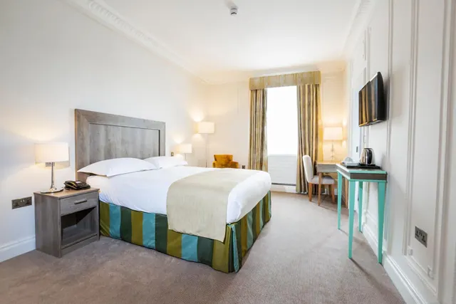 A bedroom with a made double bed, a wall-mounted television, a small desk and chair by the window, an armchair, and a kettle on a teal table.