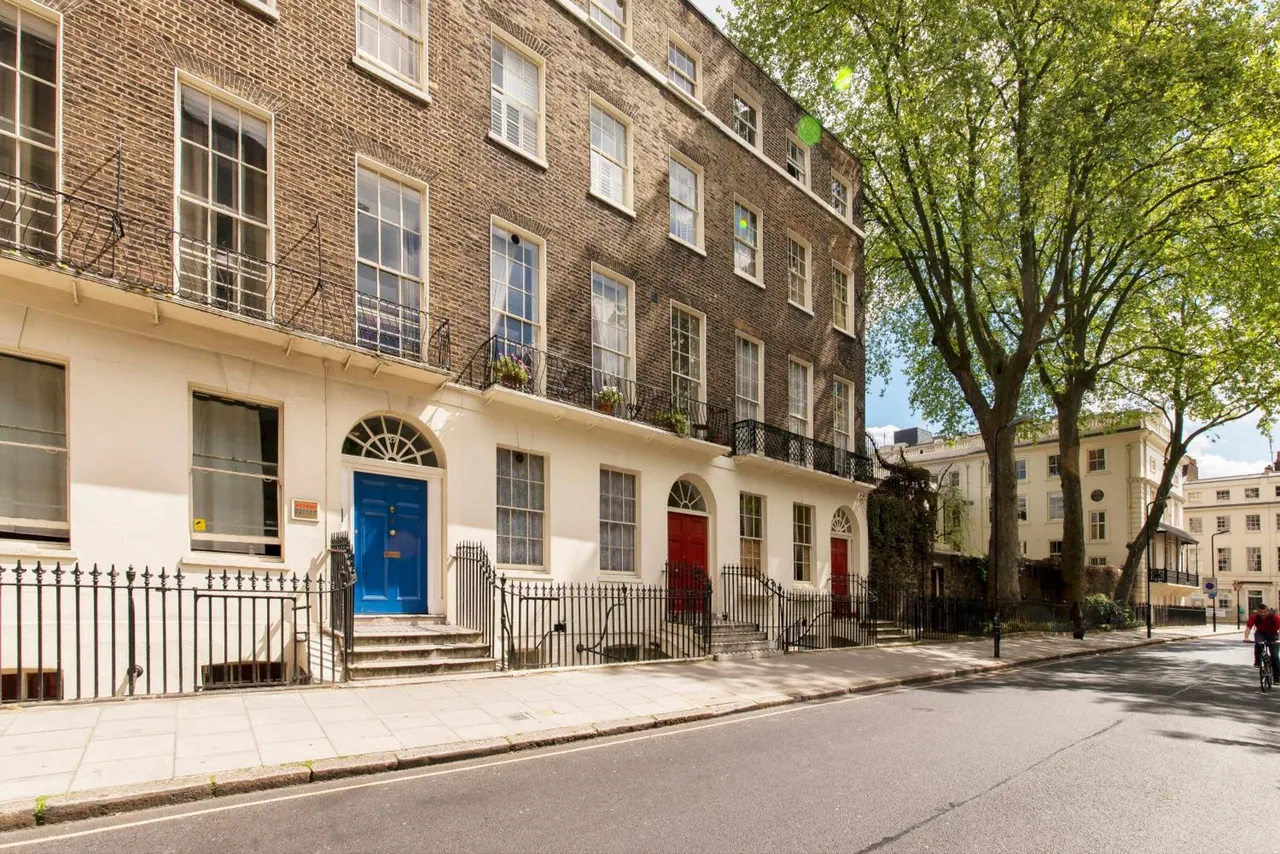 The facade of a brick Georgian terrace with a cream ground floor, blue and red front doors, tree-lined pavement and a cyclist on the road.