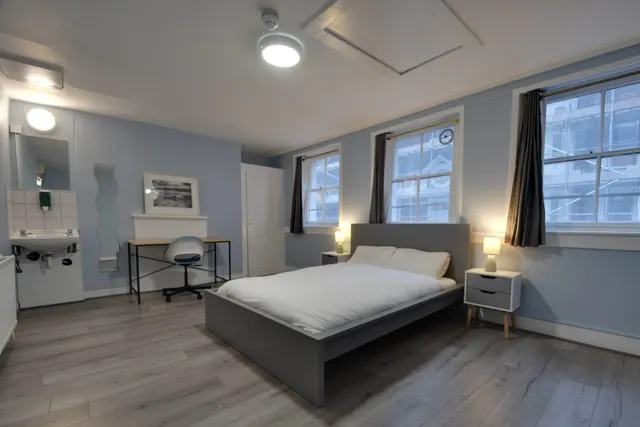 A bedroom with a double bed, two bedside tables with lamps, a work desk and chair, and a washbasin, with windows looking onto a building under scaffolding.