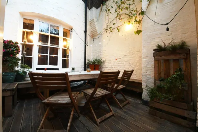 A walled courtyard terrace with a wooden deck, folding wooden chairs and a table, string lights, and potted plants beside a sash window.