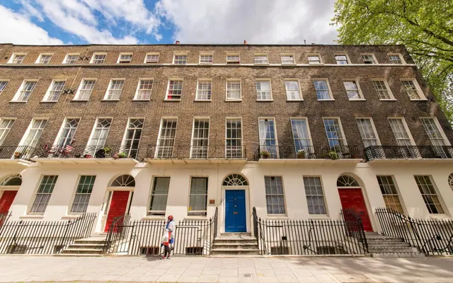 A terrace of brick Georgian houses with white ground floors, black railings, red and blue front doors, and one pedestrian walking along the pavement.