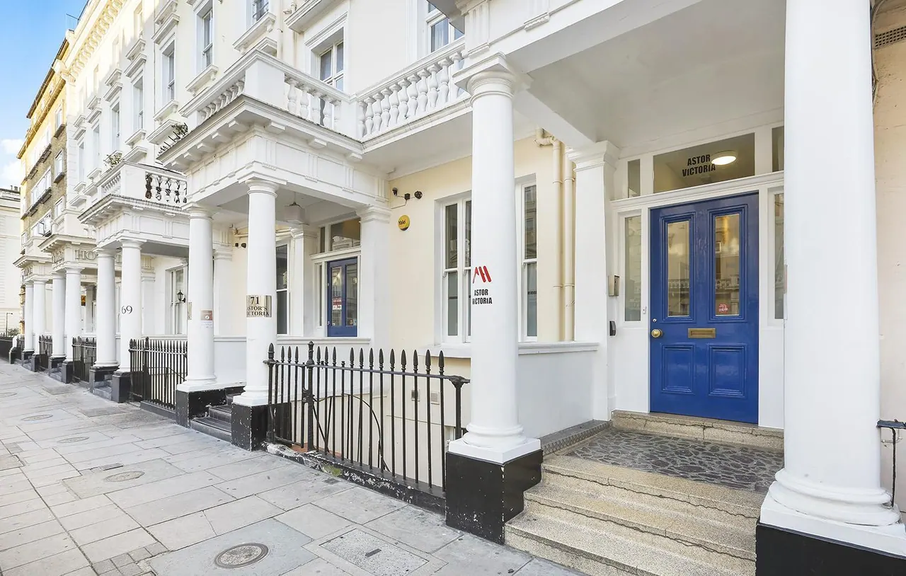 The white stucco facade of a Victorian terraced building shows a blue front door, white columns, black iron railings and Astor Victoria signage at street level.