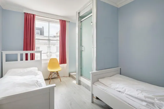 A bedroom with two single white beds, a yellow chair, red curtains at a window looking onto a building across the street, and a glass-fronted shower enclosure.