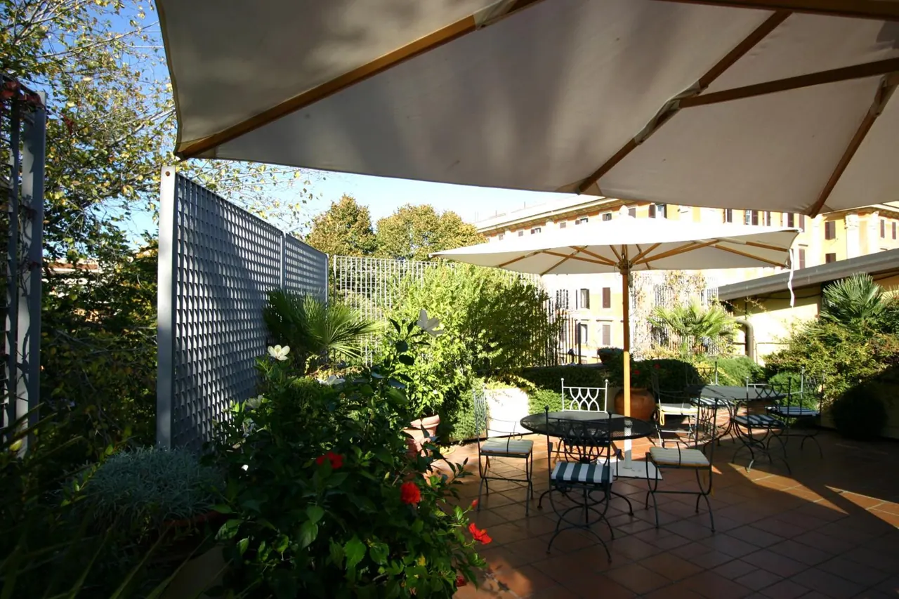 A sunlit terrace with wrought-iron tables and chairs under cream umbrellas, surrounded by potted plants, a lattice screen and a building behind.