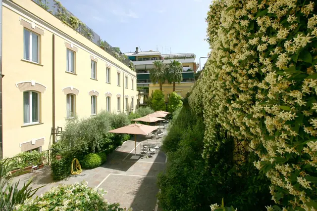 A paved courtyard with pink parasols over tables and chairs, flanked by a tall jasmine-covered wall and a yellow building.