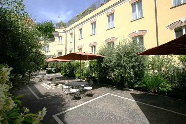A paved courtyard garden with metal tables and chairs under brown parasols, a stone fountain, olive trees and a yellow building behind.