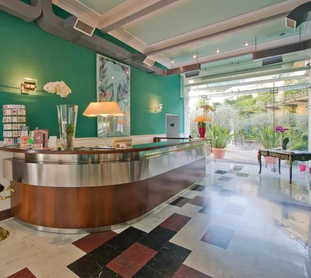 The lobby reception has a curved wood and stainless steel desk, green walls, a checkered marble floor, and glass walls facing a garden terrace.