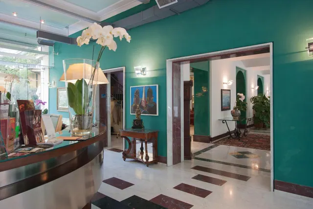 A hotel lobby with teal walls, a curved reception desk with a white orchid arrangement, a marble floor and open doorways leading to corridors.