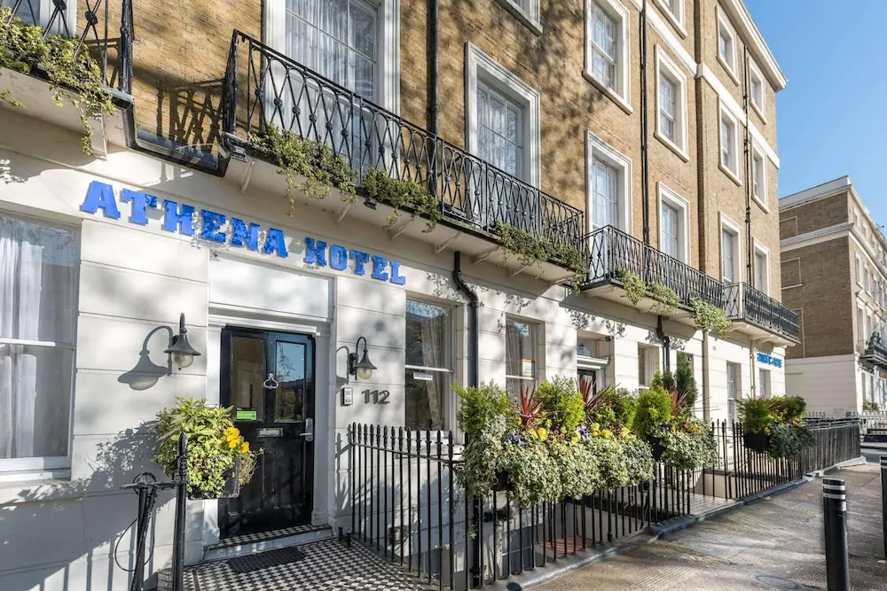 Street view of the front of a hotel with blue lettering reading Athema Hotel on a white-painted ground floor, a black entrance door, black iron balconies and flower planters on a brick terrace building.
