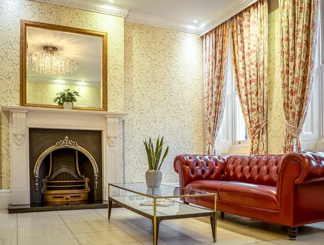 A lounge area with a marble fireplace, a gilt-framed mirror, floral curtains, a red leather Chesterfield sofa and a glass coffee table.