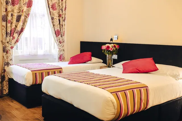 A bedroom with two beds, red cushions, floral curtains at the window and a vase of roses on the bedside table.