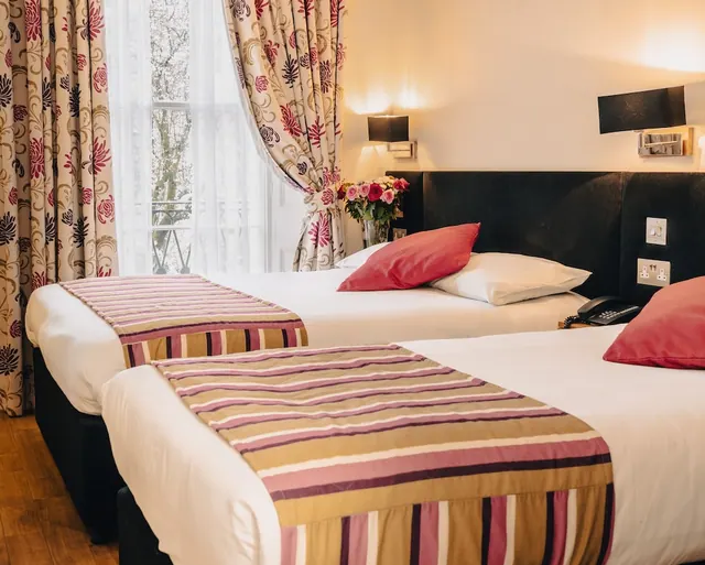 A hotel bedroom with two beds side by side, striped throws, red and white pillows, floral curtains and a window with sheer net curtains.