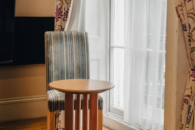 A striped upholstered chair and a round wooden side table stand beside a window with sheer white curtains and patterned drapes.