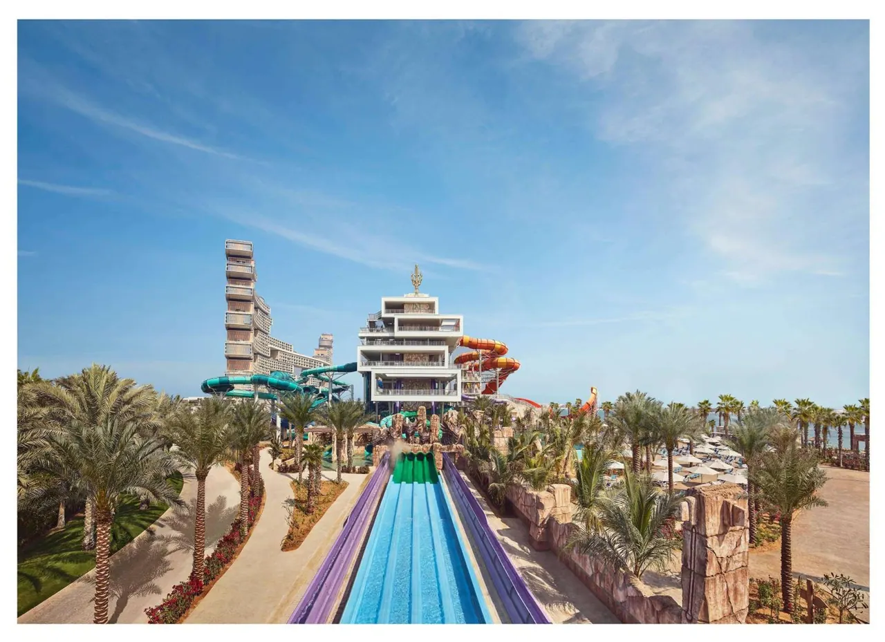 A water park with a long blue water slide running toward a white multi-storey building with spiral slides, palm trees on both sides, and white umbrellas on the right under a clear blue sky.