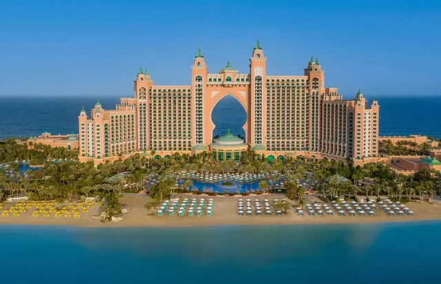 Aerial view of a large pink hotel building with a central arch, beside a sandy beach with rows of sun loungers and umbrellas and the sea beyond.