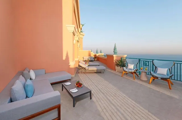 A furnished terrace with a sofa, a coffee table with a fruit bowl, lounge chairs and a view over the sea.