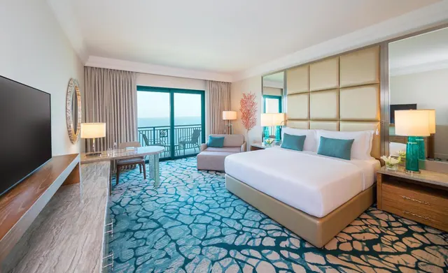 A bedroom with a large white bed, a teal patterned carpet, a seating area, a wall-mounted television and sliding doors opening onto a balcony with a view of the sea.