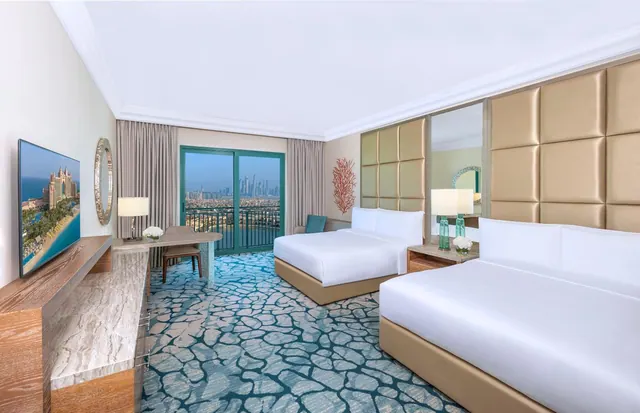 A hotel bedroom with two beds, a desk, a wall-mounted television and a glass door opening onto a balcony with a city skyline beyond.
