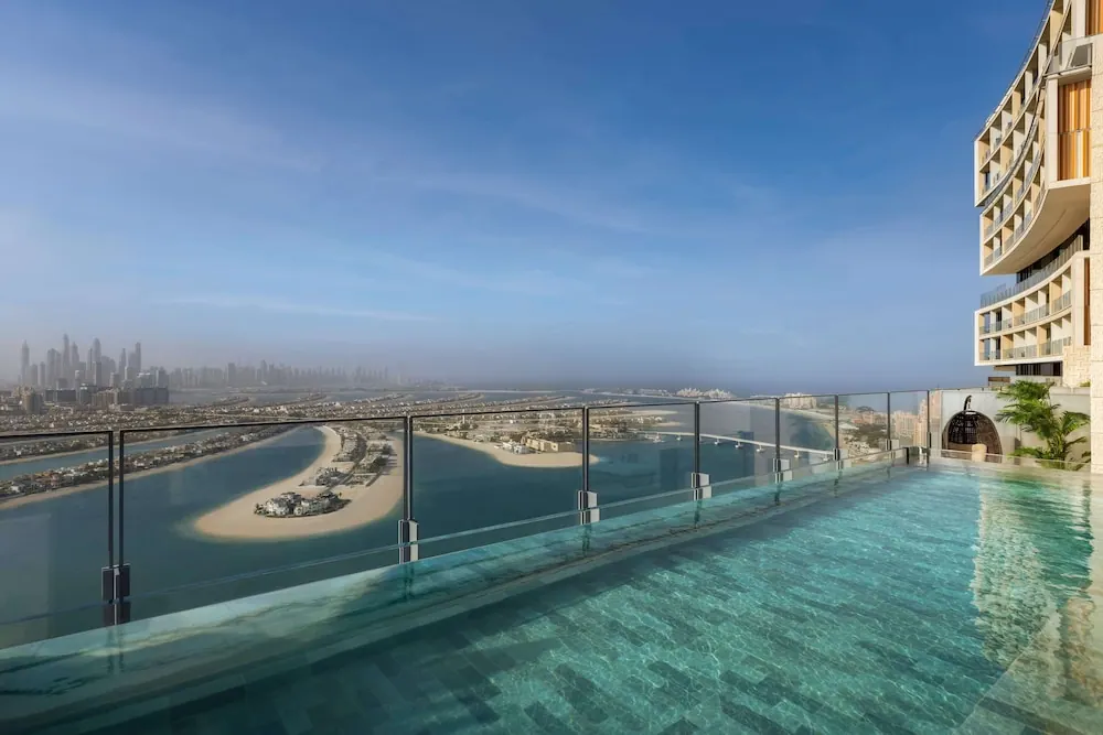 An infinity edge rooftop pool with a glass railing overlooks the sea, a crescent-shaped palm island and a distant city skyline under a clear blue sky.