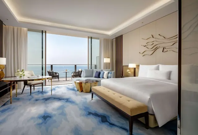 A bedroom with a king bed, a seating area and a desk, with glass doors opening onto a balcony that looks out over the sea.