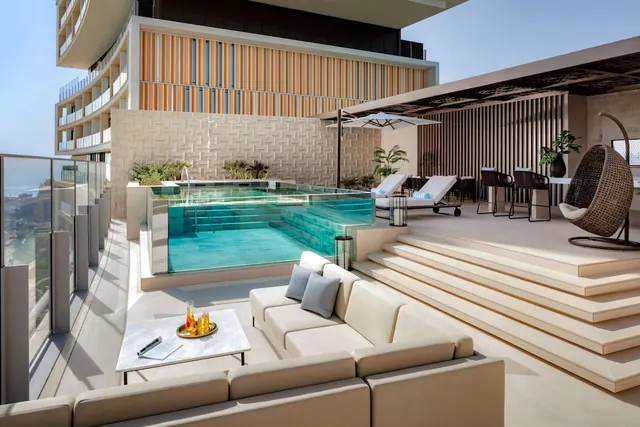 A rooftop terrace with a small glass-edged pool, sofa seating, sun loungers, a hanging chair and a strip of sea visible at the left edge.
