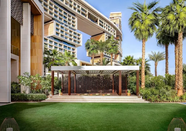 A green lawn leads to a covered wooden pavilion with steps, with tall palm trees and a pale multi-storey hotel building behind it.