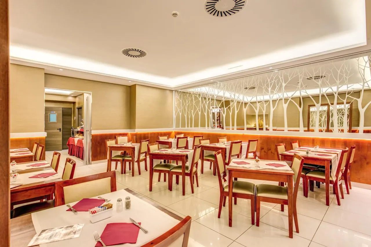 A restaurant dining room with set tables, red napkins, wooden chairs, and a mirrored wall with white tree decals.
