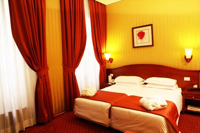 A bedroom with two beds side by side under a wooden headboard, red curtains at the windows, yellow striped walls and a framed rose picture.