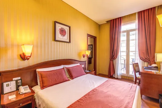 A double bed with red pillows and a red throw sits in a yellow-striped bedroom beside a desk and tall doors opening onto a balcony.
