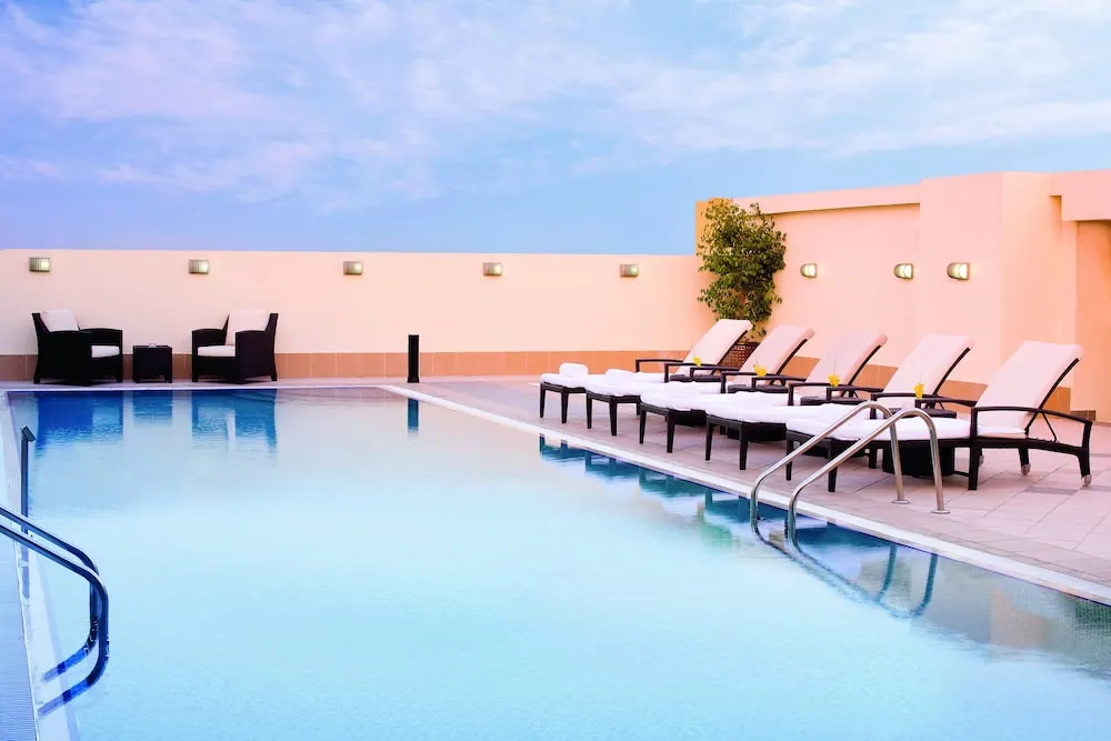 A rooftop swimming pool with white sun loungers and dark armchairs beside a peach-coloured wall under a pale blue sky.