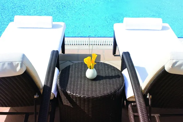 Two white sun loungers with towel rolls sit beside a wicker side table holding a vase of yellow flowers at the edge of a blue swimming pool.
