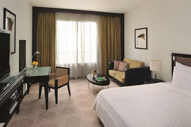 A hotel bedroom with a king bed, a sofa and armchair, a desk with a chair, and a television, with sheer curtains over a window.