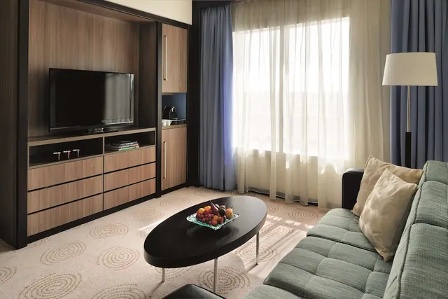 A hotel living area with a wall-mounted television in a wooden cabinet, a grey sofa, a dark oval coffee table holding a fruit bowl, and a floor lamp beside sheer curtains.