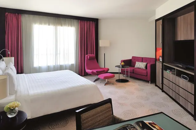 A hotel bedroom with a made king bed, a pink armchair with ottoman and a pink sofa by a round side table, and a wall unit holding a flat-screen TV.