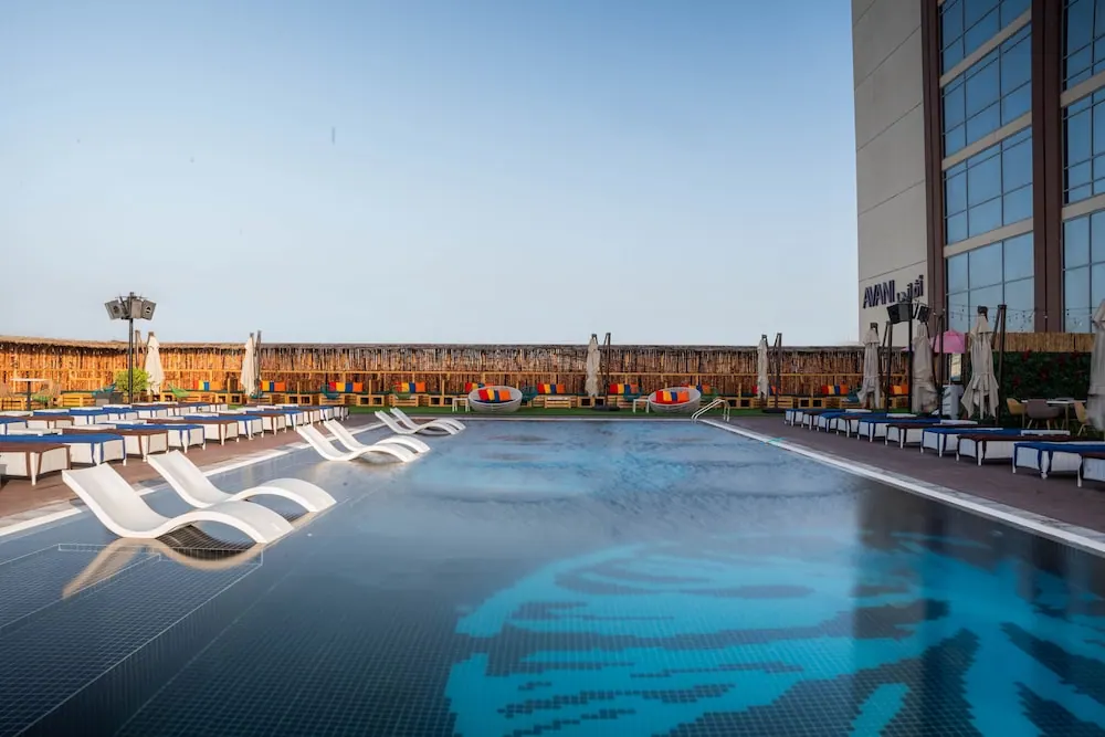 An outdoor pool with white sun loungers in the water and blue-cushioned loungers along the deck, a reed fence behind, and a glass building with an Avani sign to the right.