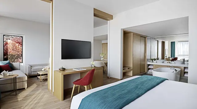 A hotel suite bedroom with a large bed, wall-mounted television, desk with red chair, lounge seating, and a freestanding bathtub and double vanity visible through glass in the bathroom.