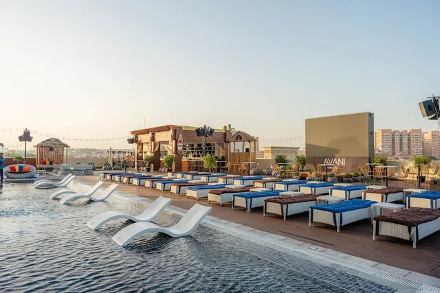 A rooftop pool with white submerged loungers, rows of blue-cushioned sun loungers on the deck, a bar pavilion, and residential towers in the background.