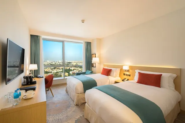 A twin-bedded hotel room with a wall-mounted television, a desk with a coffee machine, and a large window overlooking a city with low buildings and greenery.