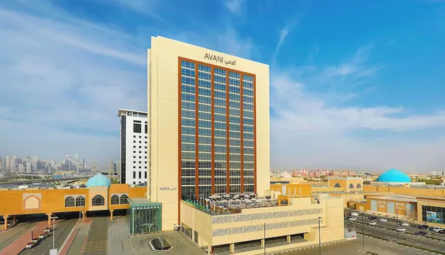 Elevated view of the AVANI hotel tower with a rooftop terrace under umbrellas, adjacent low buildings, a parking area and a city skyline in the background.