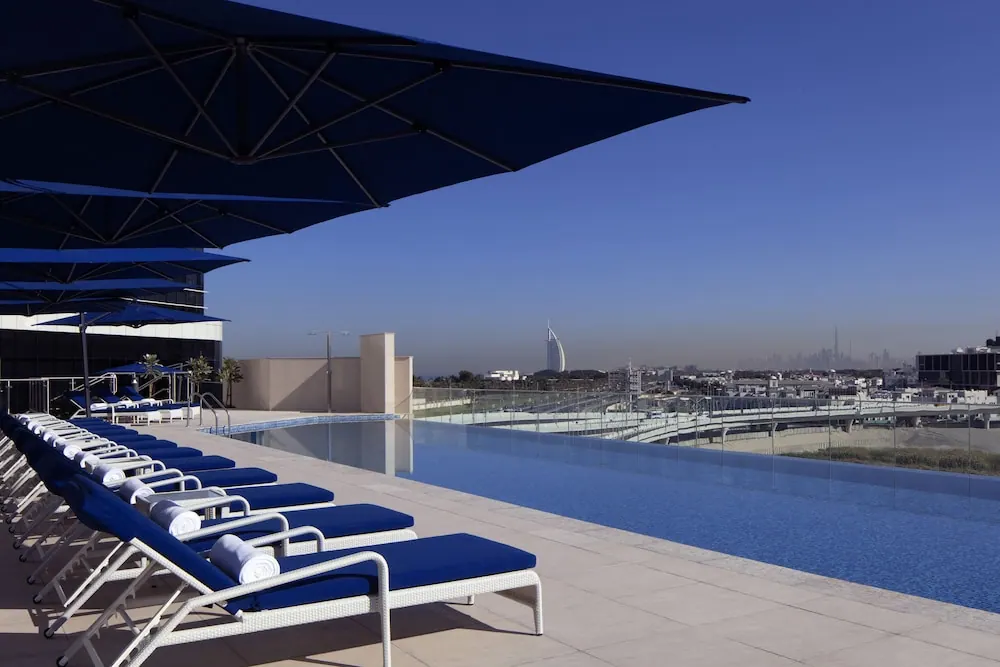 An infinity-edge rooftop pool with white and blue loungers and blue umbrellas, overlooking a hazy view of the Dubai skyline with Burj Al Arab in the distance.