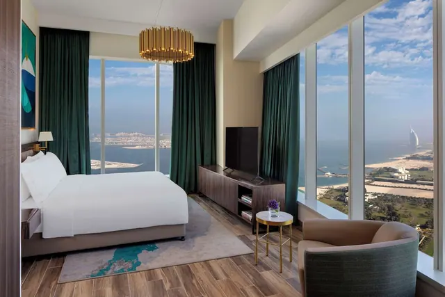 A bedroom with a king bed, a television on a wooden console, an armchair, and floor-to-ceiling windows looking over the sea toward Burj Al Arab.