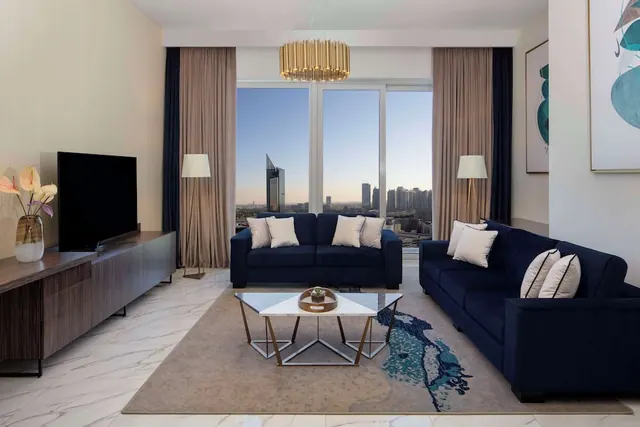 A living room with two navy sofas, a wall-mounted television, and floor-to-ceiling windows showing a city skyline.