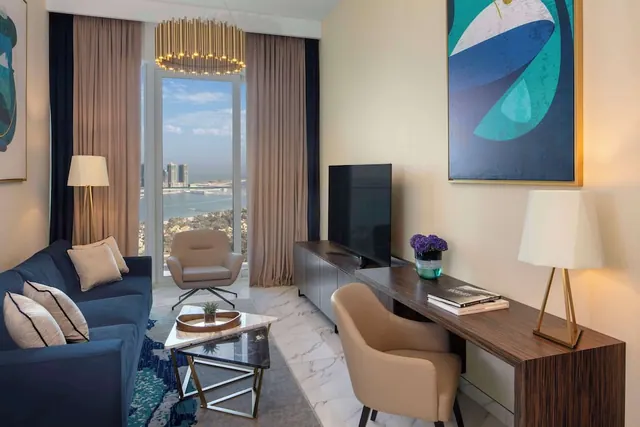 A living room with a navy sofa, a desk and television, and a floor-to-ceiling window showing the sea and distant towers.