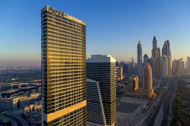Aerial view of a gold-glass high-rise with the Avani name on its roof, with other towers, low-rise buildings and a multi-lane road visible at sunset.