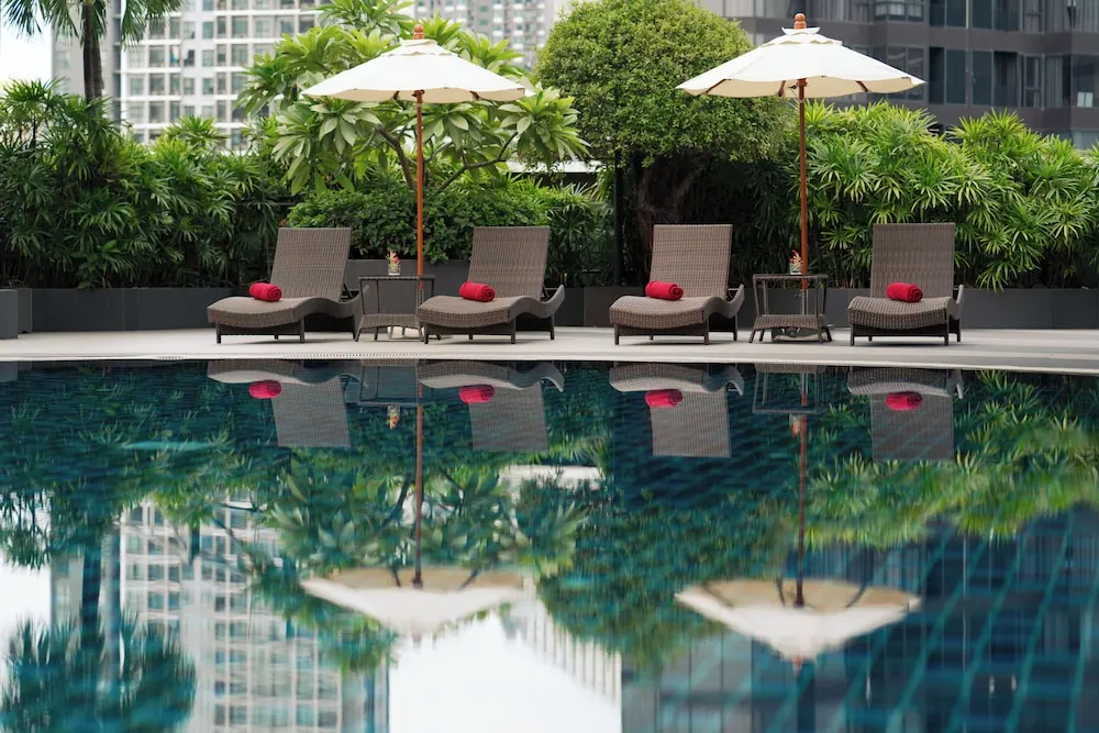 Four wicker sun loungers with red towels and two cream parasol umbrellas stand beside a pool, with tall buildings and greenery reflected in the water.