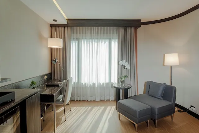 A hotel room living area with a wooden desk and chair on the left, a grey armchair with ottoman beside a round side table, and a window hung with sheer curtains.