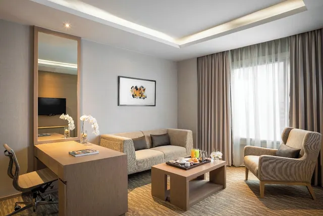 A hotel room living area with a sofa, armchair, coffee table holding a breakfast tray, and a desk beside a mirror, with a television visible in the mirror's reflection.