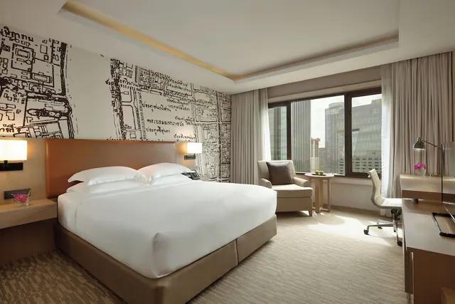 A bedroom with a king bed, an armchair beside a window overlooking city towers, and a work desk on the right.