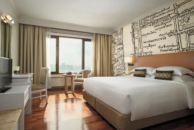 A bedroom with a large white king bed, a desk and television, and a window looking out over distant city buildings.